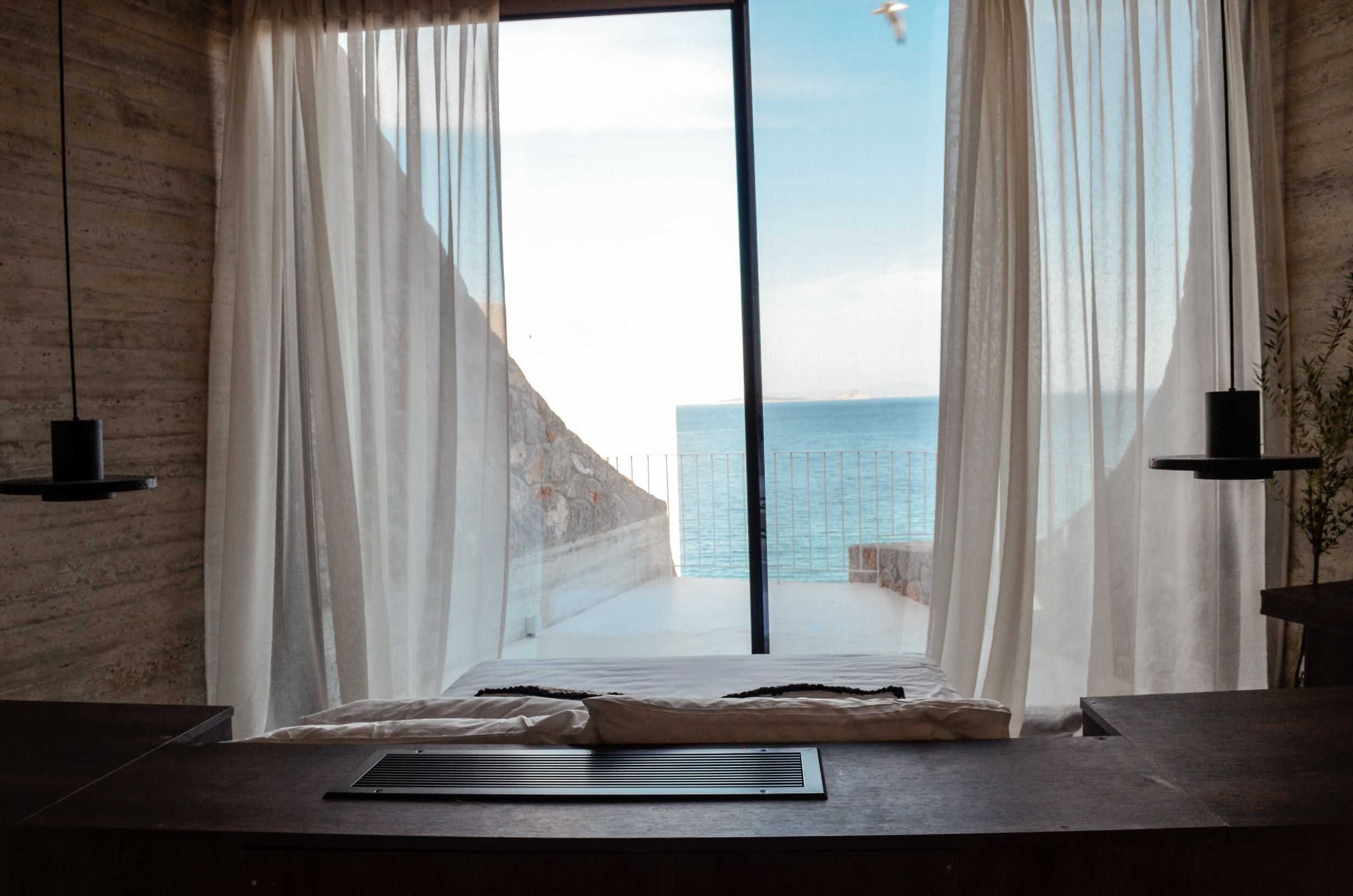 Bedroom window framing a sea view between sheer curtains — Amoni