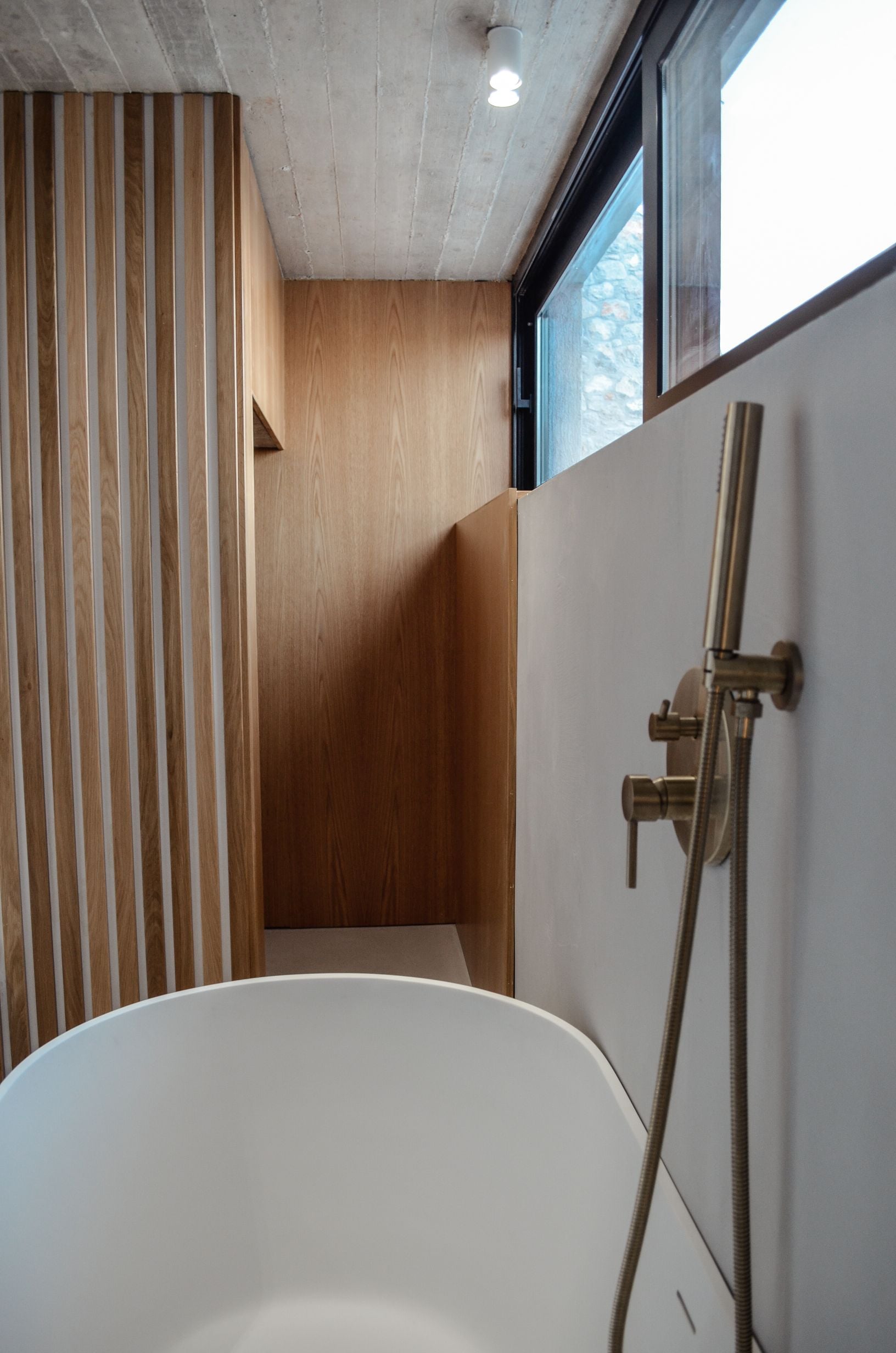 Freestanding bathtub beside a timber wall and a high window — Amoni